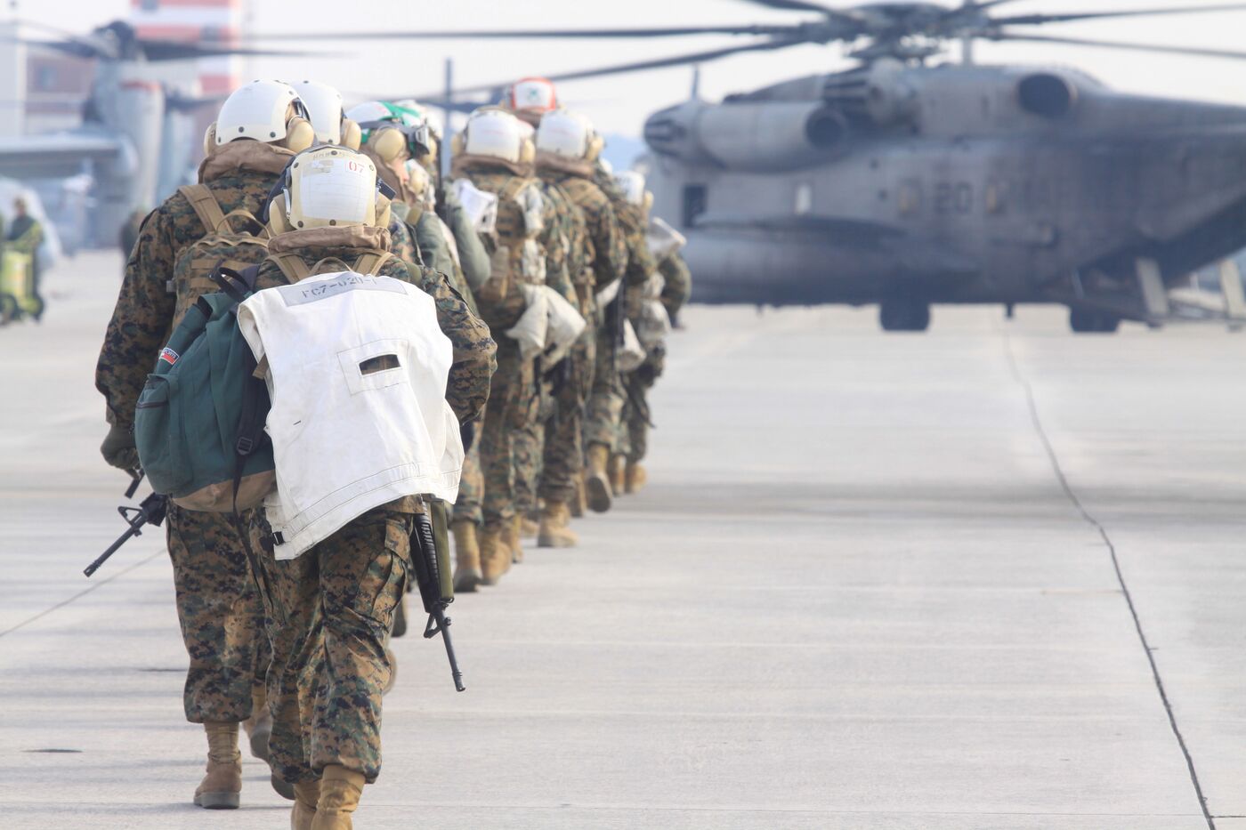 A line of Marines in flight helmets walking down a hazy flightline toward a CH-53 helicopter