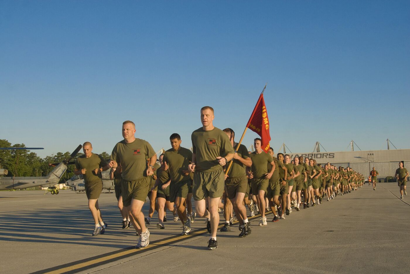 Marines running in formation across a flight line at dawn, carrying a unit flag