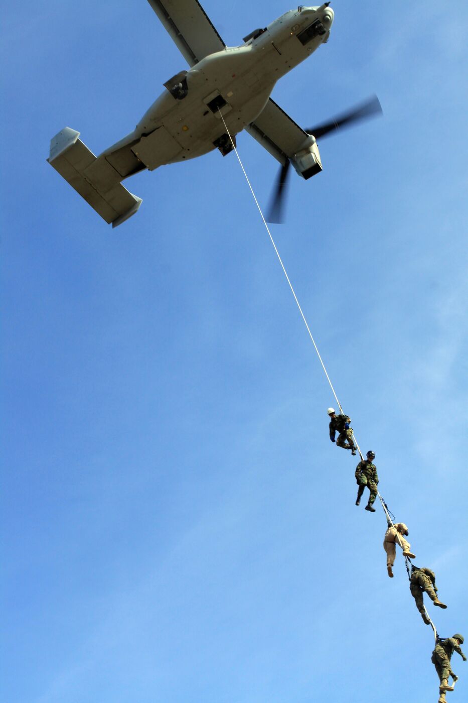 Looking straight up at an MV-22 Osprey as five Marines hang from a single rope beneath it