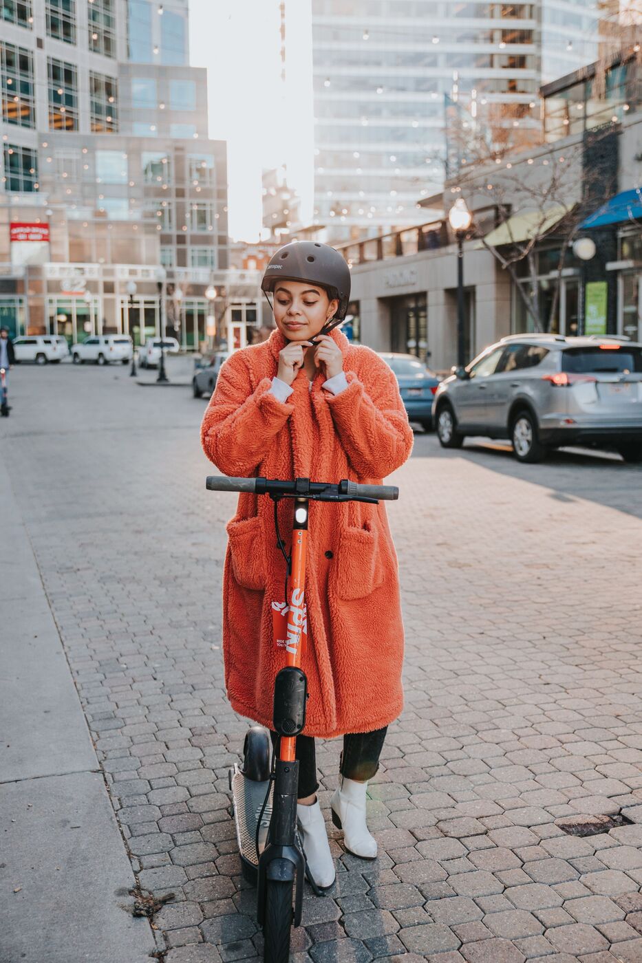 A rider in an orange coat fastening her helmet beside a Spin scooter at golden hour