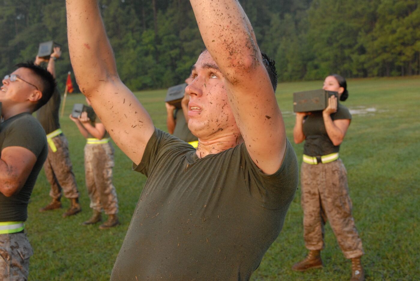 A Marine pressing an ammo can overhead on a grassy field at dawn