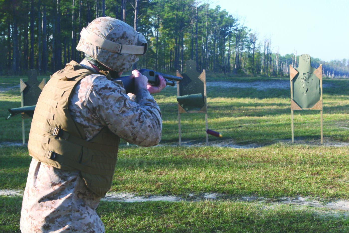 A Marine firing a shotgun on a range, a shell casing mid-air